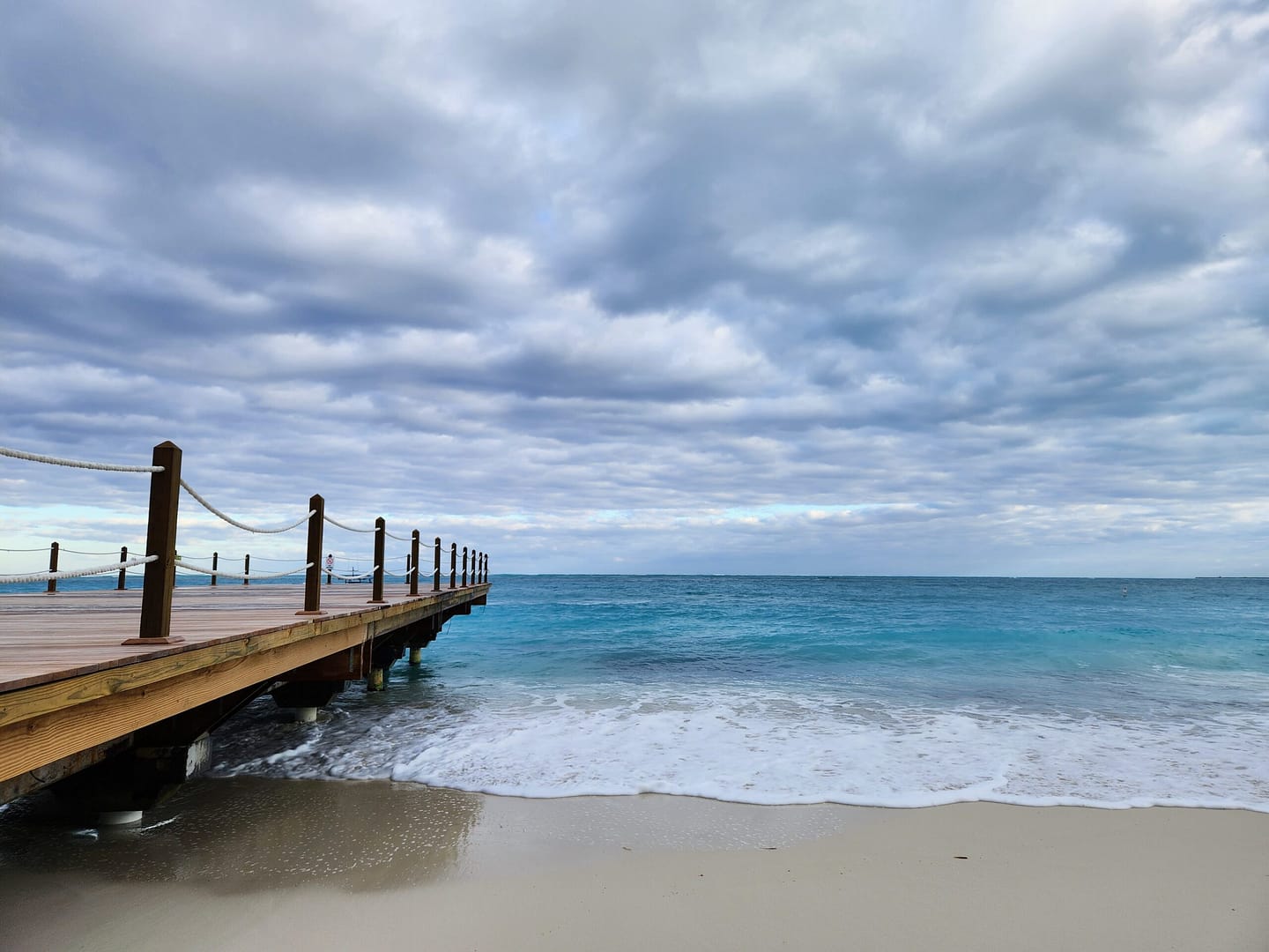 Wooden dock into the ocean on a white sandy beach with turquoise water and a moody sky