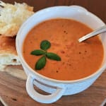 Bowl of creamy tomato basil soup, topped with fresh sprig of basil, with chunky bread sitting next to the soup bowl.