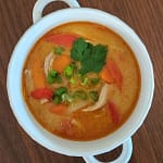 Bowl of thai chicken curry soup, including chicken, peppers, carrots, and bamboo shoots in a creamy red curry broth and topped with cilantro and green onions