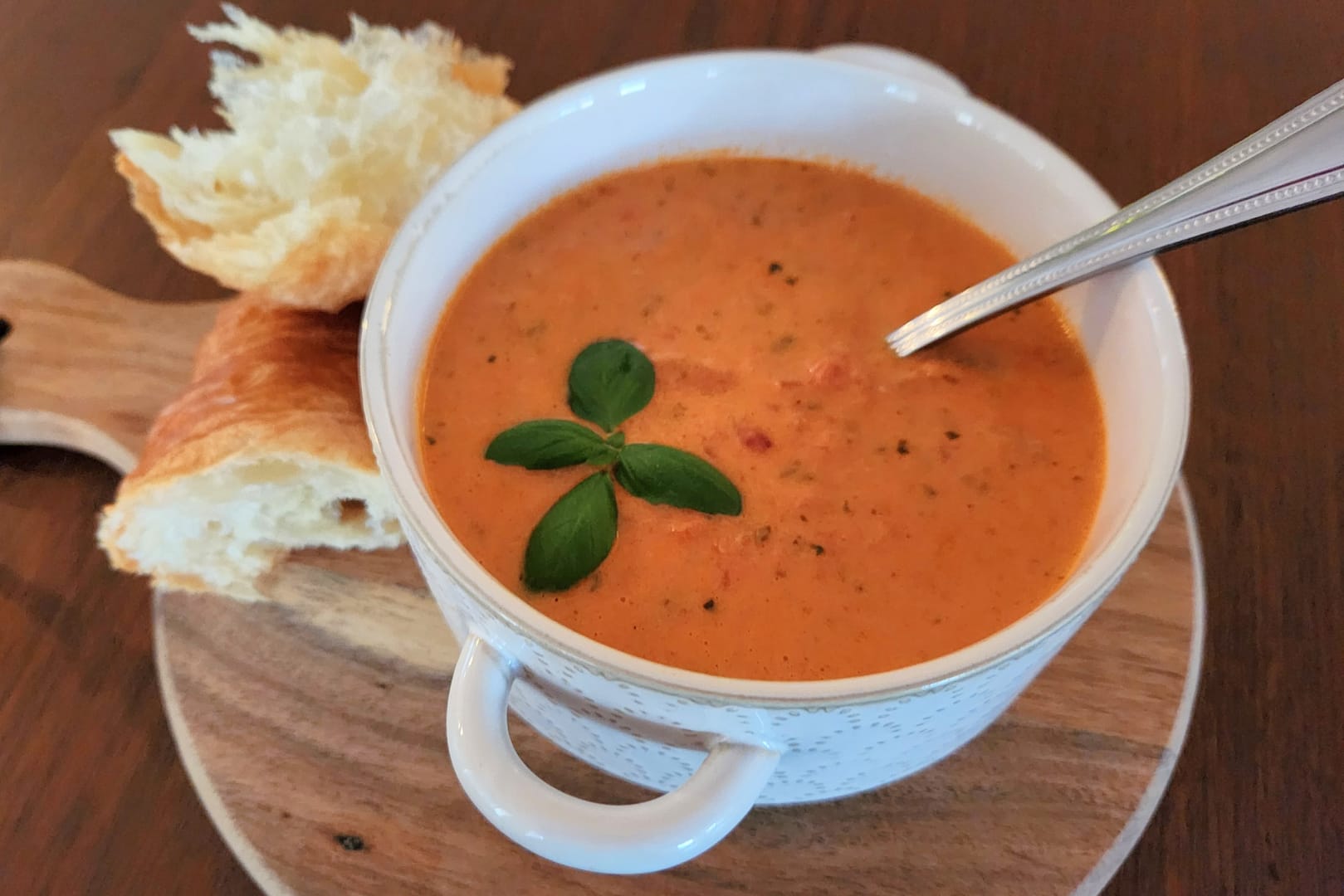 Bowl of creamy tomato basil soup, topped with fresh sprig of basil, with chunky bread sitting next to the soup bowl.
