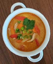 Bowl of thai chicken curry soup, including chicken, peppers, carrots, and bamboo shoots in a creamy red curry broth and topped with cilantro and green onions