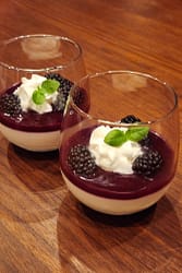 Panna cotta served in stemless wine glasses topped with blackberry sauce, whipped cream, basil sprigs, and fresh blackberries